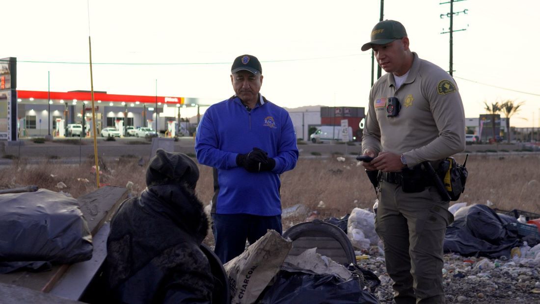 Deputy and Supervisor Joe Baca Jr speaking to a person at a homeless encampment near a gas station.