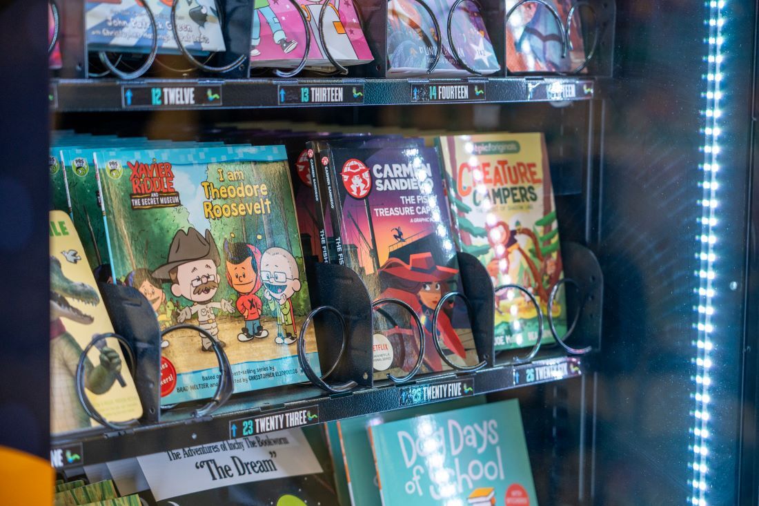 A vending machine filled with children's books, including titles like "I am Theodore Roosevelt" and "Creature Campers," displayed on multiple shelves.