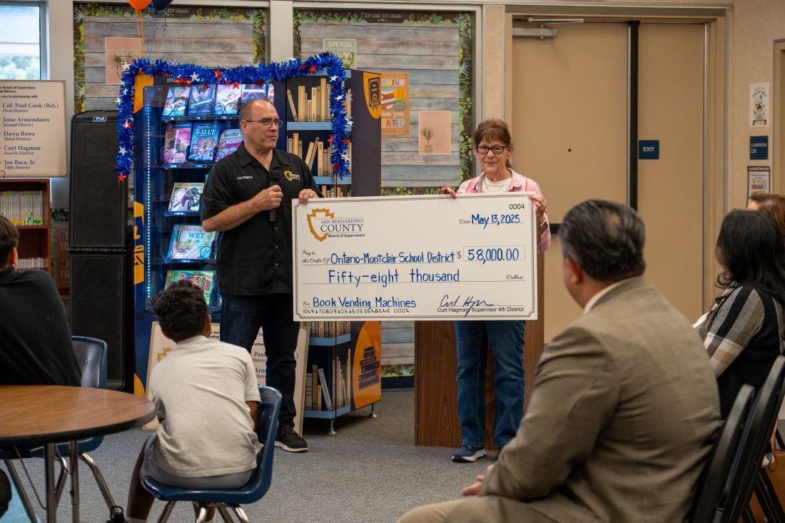 A man and a woman hold a large check for $58,000 made out to Ontario-Montclair School District for book vending machines, while an audience watches.