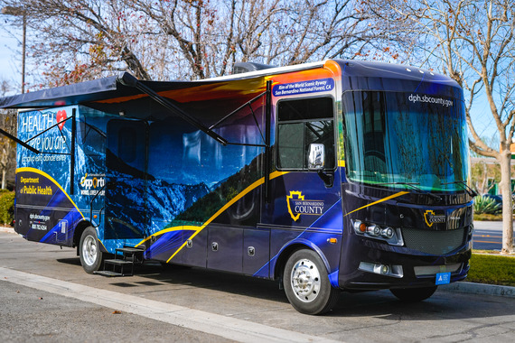 A blue mobile health vehicle from the San Bernardino County Department of Public Health is parked on a sunny day.