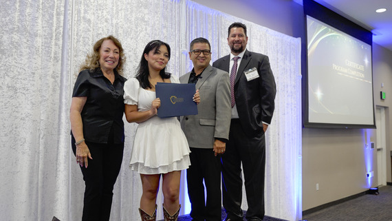A young woman holds a county certificate while posing with three county officials against a curtain backdrop, with a video play button overlay.