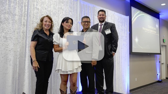 A young woman holds a certificate while posing with three officials against a curtain backdrop, with a video play button overlay.