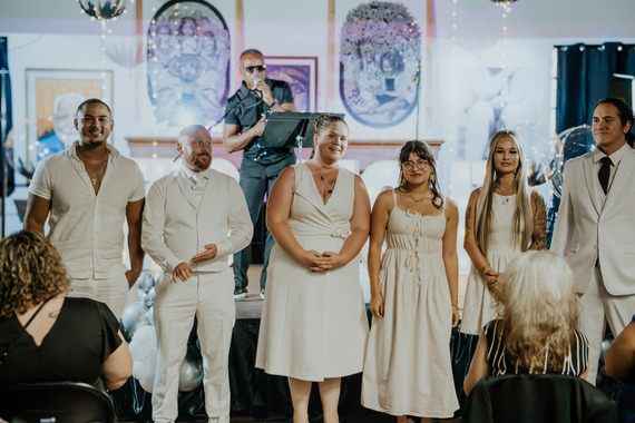 Six people stands at the front of a room with onlookers seated around them and an artist performs in the background. 