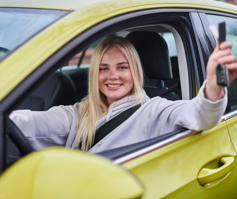 A blonde teenage girl sits behind the wheel of a green car, smiling and holding something in her left hand. 