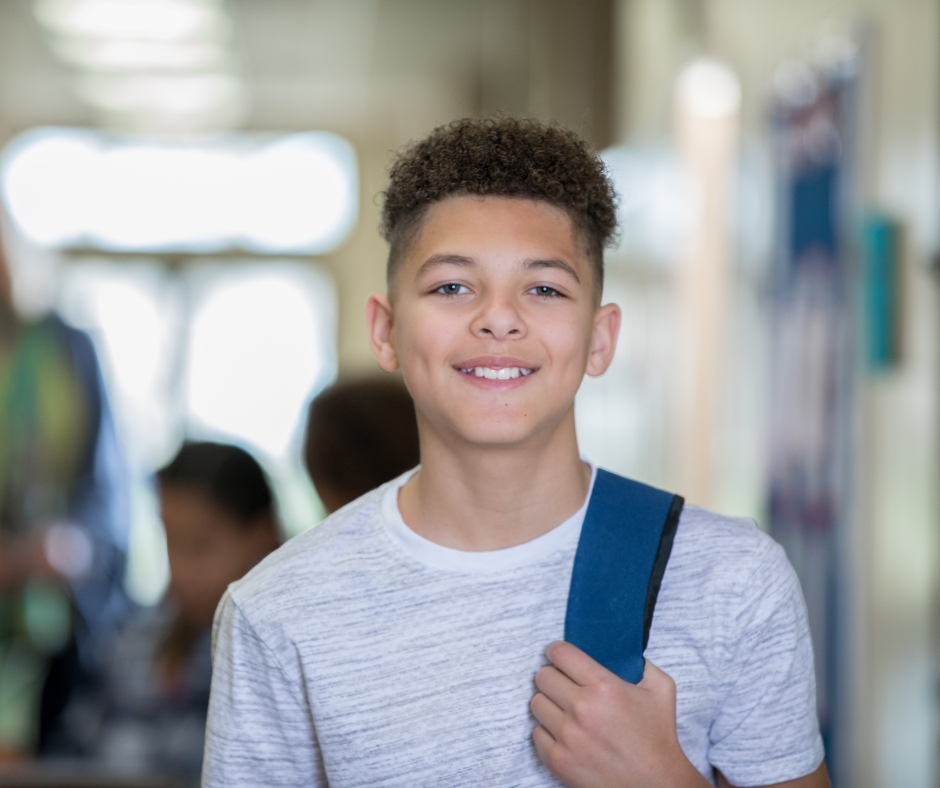 A young male student with curly hair and a backpack slung over one shoulder stands smiling in the hallway of a school