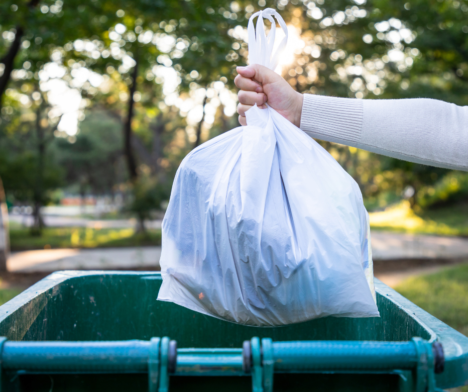 A person’s hand holding a white garbage bag over a green outdoor trash bin in a park setting with trees and sunlight in the background.
