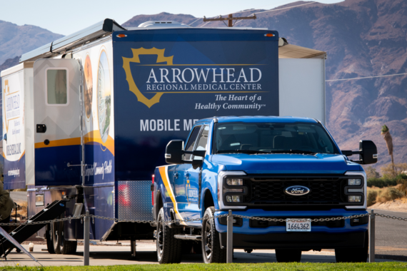 A blue Ford truck is parked in front of a large mobile medical clinic trailer from Arrowhead Regional Medical Center.