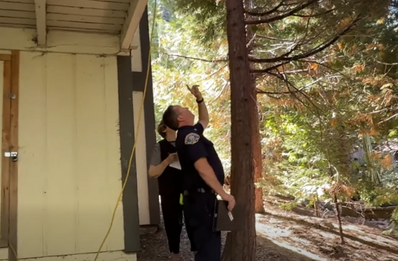 Fire department officer conducts a home wildfire safety assessment, pointing toward tree branches near the roofline. Another person takes notes.