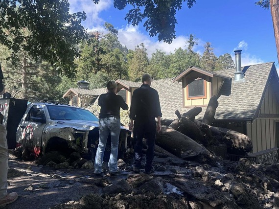 Two people stand near a mud-covered truck and fallen trees in front of a damaged house after a mudslide in a forested area