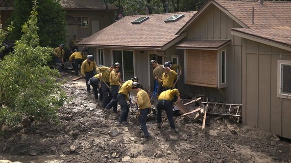 About a dozen San Bernardino County firefighters work with shovels to remove mud from around a home in Forest Falls