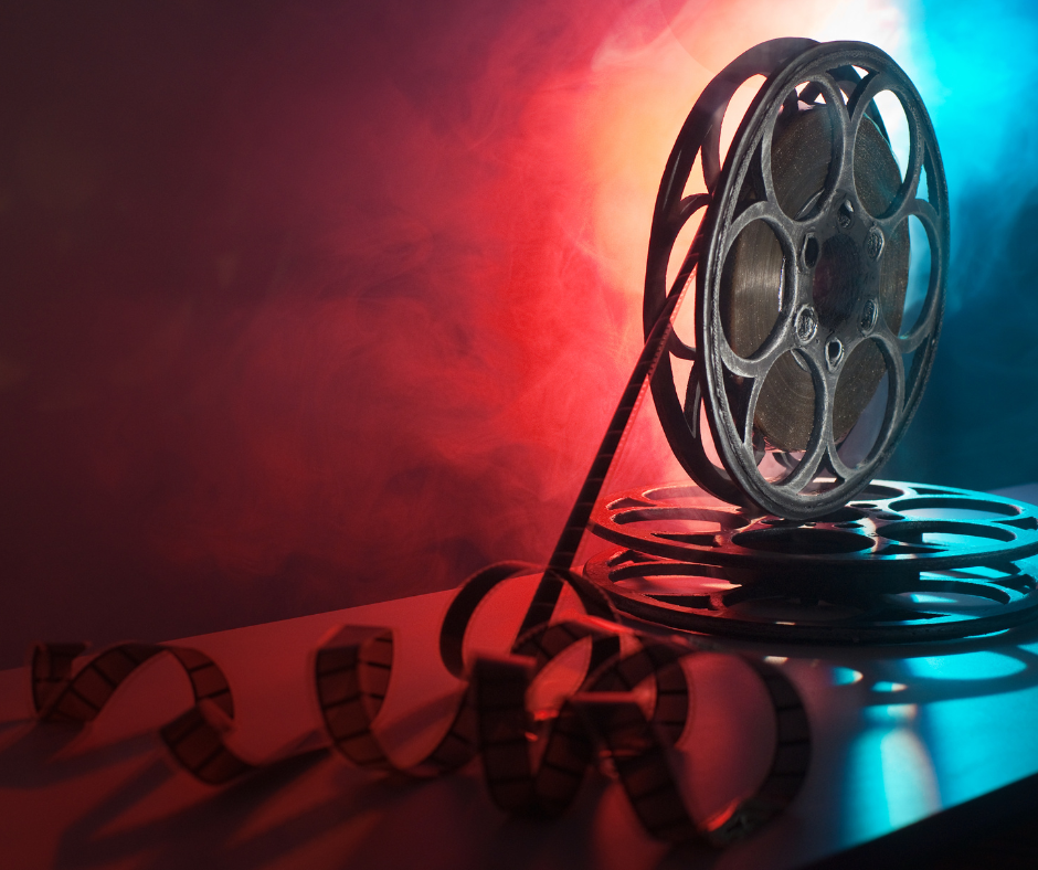 A reel of movie film sits on a table, slightly unrolled, against a dark, noir-ish background and scene.