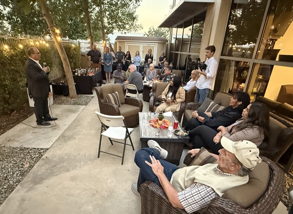 A group of people seated and standing outdoors, listening to a speaker during an evening event with string lights.