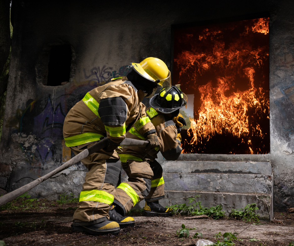 Two firefighters in full turnout gear hold a hose as they work to put out a fire burning in a house.