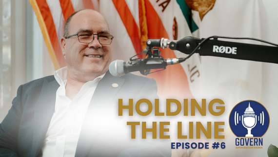 Supervisor Hagman speaks into a podcast mic with flags behind him for Episode 6.