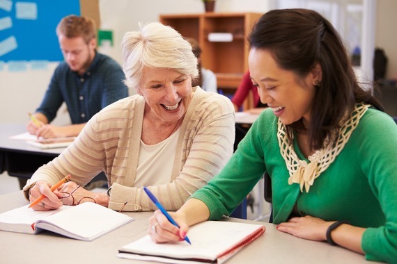 Two adults smiling and writing together in a classroom setting during a learning session.
