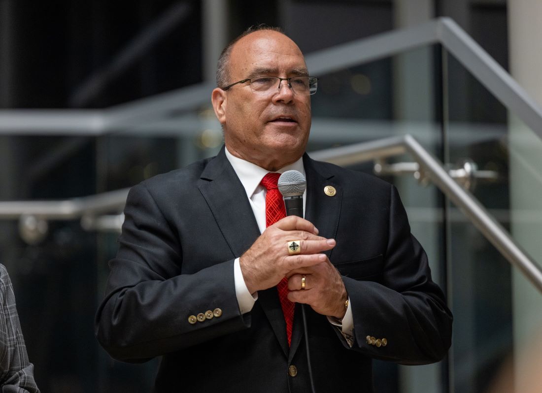 Man in suit holding a microphone and speaking during an indoor community event.