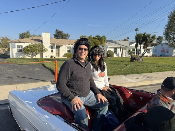 Two people wearing winter hats sit in a convertible during a sunny neighborhood parade.