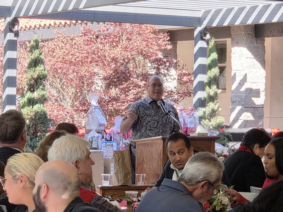 Man speaking at a podium during a festive outdoor gathering with seated attendees listening.