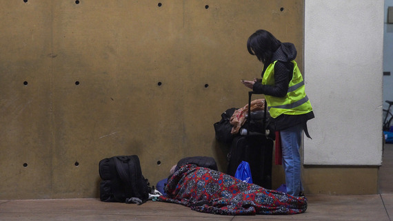 A person in a high visibility vest using a smartphone stands next to a person lying on the ground under a colorful blanket. 