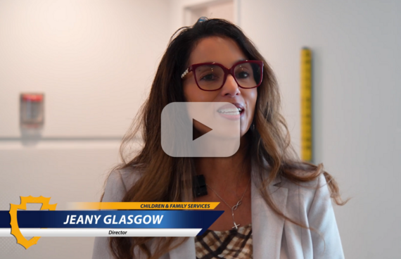  Jeany Glasgow, Director of Children & Family Services, speaks indoors, wearing red glasses and a light blazer with a microphone clipped on. 