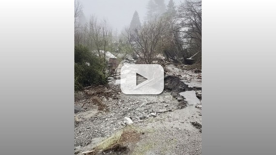 Storm runoff and debris cover a washed-out roadway, with broken pavement, rocks, and fallen branches amid trees under foggy conditions. 