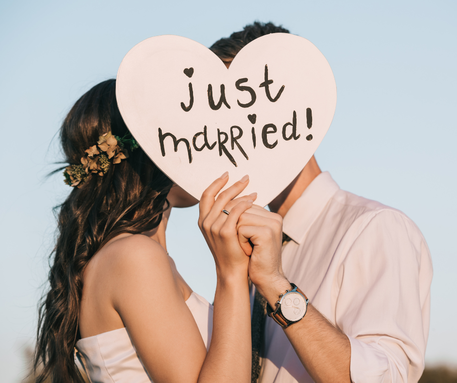 A newly married couple holds a heart-shaped sign that says "just married!" while kissing. One person wears a floral crown, and the other has a watch.