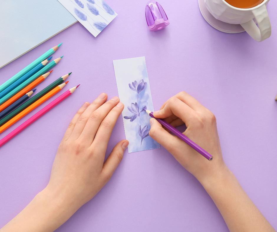 Hands coloring a handmade bookmark with soft purple flowers on light blue paper. Colored pencils, a notebook, and a cup of tea sit on a lavender desk