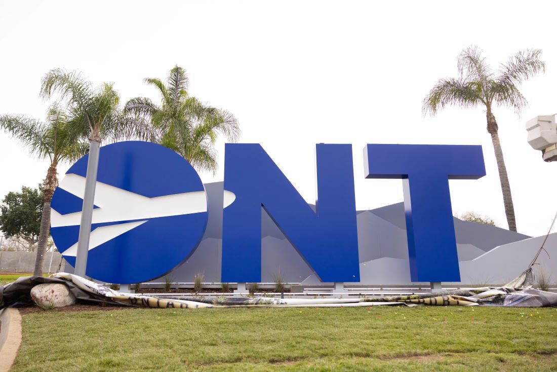 Large blue "ONT" monument sign with mountains and palm trees in the background and grass in front. 