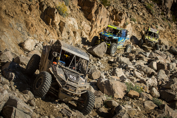 Off-road race vehicles on a rocky desert trail during an extreme rock-crawling competition in a rugged canyon landscape.
