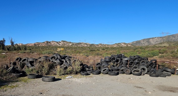 Large piles of tires dumped on a dirt road in a mountain area under a blue sky, with low vegetation, a fence line, and power lines.