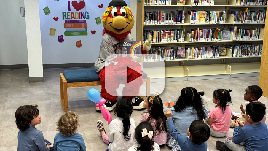 A colorful mascot reads to young children sitting on the floor in a library. Bookshelves are in the background. The mood is joyful and engaging.
