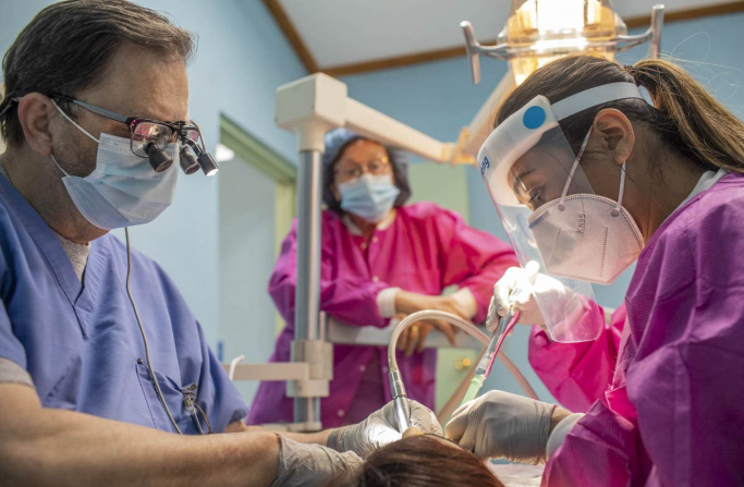 Two doctors in full protective gear work on a young dental patient in a chair as an assistant looks on from a light post.