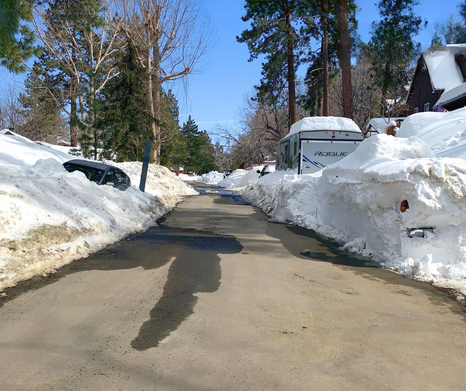 Snow-lined mountain street with car part buried in snow & a white RV trailer parked on the right, surrounded by deep snow under a blue sky.