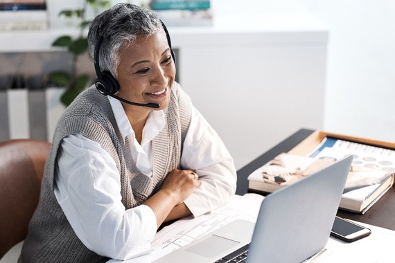 Person speaking with a woman on a computer, white cell phone top of a wooden desk, blurred background.