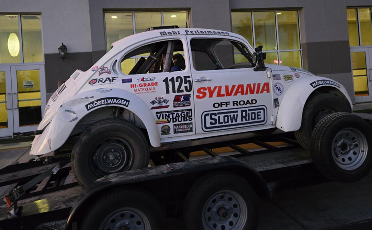 White Class 11 Volkswagen Beetle race car mounted on a flatbed trailer outside a building at dusk. Race number 1125 displayed on the door.