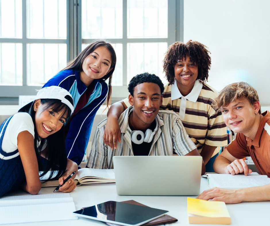 Five diverse young adults smile at the camera while gathered around a table with a laptop, notebooks, and a tablet in a bright room with large windows