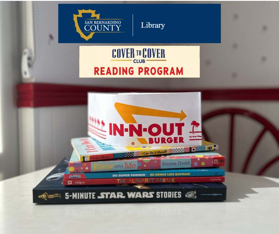 In-N-Out Burger hat resting on a stack of children’s books beneath a San Bernardino County Library “Cover to Cover Reading Program” banner.
