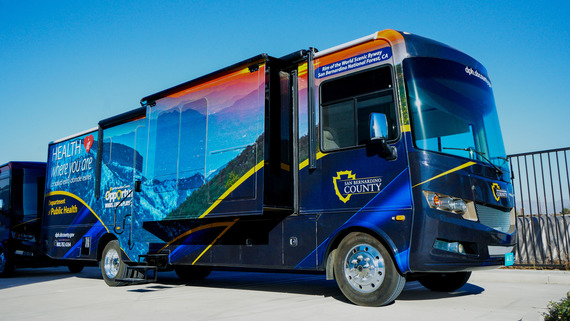 San Bernardino County mobile health clinic bus with colorful graphics and “Health where you are” branding logos parked outdoors.