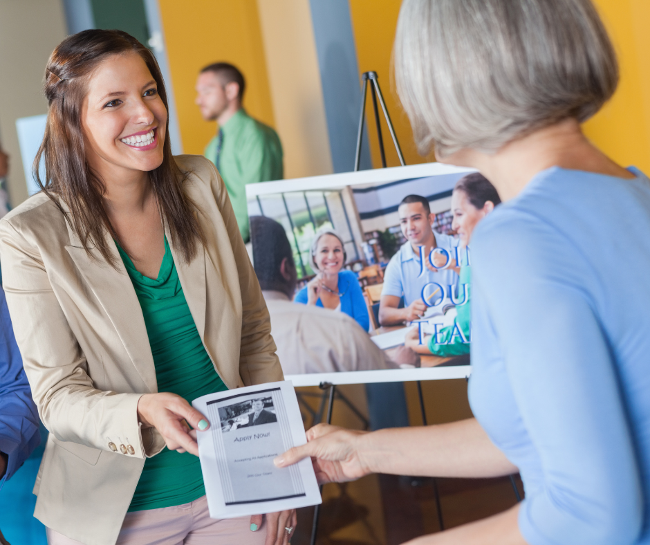 woman at a job fair smiles and hands a flyer to another attendee, with a recruitment display and people networking in the background.