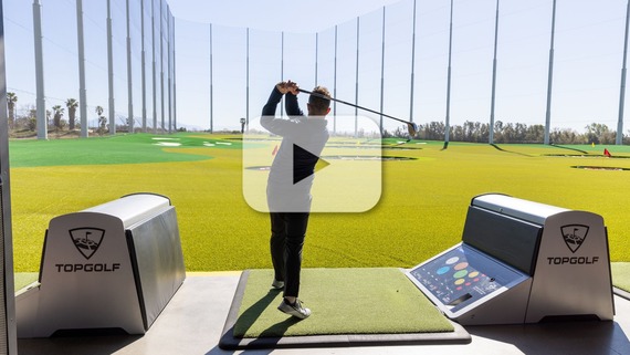 Person swinging a golf club at a Topgolf driving range, viewed from behind, with green targets and tall netting, and a video overlay button.