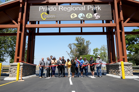 People stand behind a red ribbon under a large Prado Regional Park sign with the County logo and hiking, fishing, bicycling and camping icons.