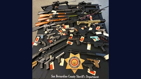 Table with rifles, shotguns, and handguns tagged as evidence; Sheriff-Coroner San Bernardino County badge, San Bernardino County Sheriff's Department.