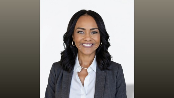 Smiling woman in gray blazer and white blouse, hoop earrings and necklace, against light background with dark border on sides.