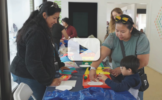 Two women assist a child with crafts at a table with colorful materials; other individuals in the background; video play button overlay.