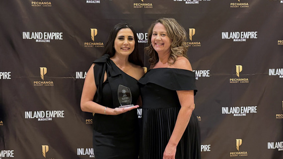 Two women in elegant black dresses pose with an award against a backdrop featuring "Inland Empire Magazine" and "Pechanga Resort Casino" logos. 