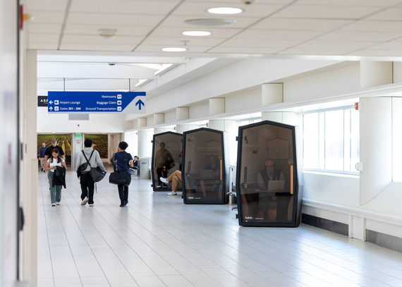 Travelers walk through an airport terminal as passengers use Kabin work booths along the wall for quiet workspace and meetings.