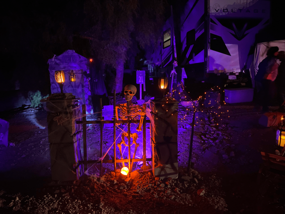 Halloween campsite decorated with glowing skeleton behind fence surrounded by purple & orange lights lanterns & spooky props near an RV at night.
