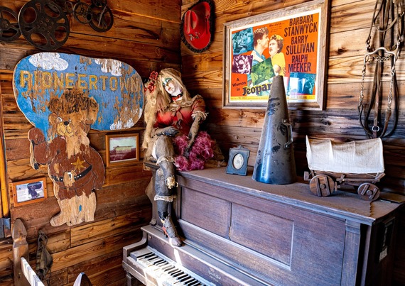 A wooden room with a vintage piano and antique doll seated on top. The walls display various items, including a cowboy-themed "Pioneertown" sign.