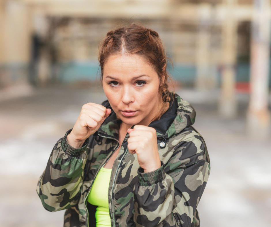 woman in camouflage jacket stands in defensive boxing stance w/fists raised looking directly at camera in indoor setting w/blurred background.