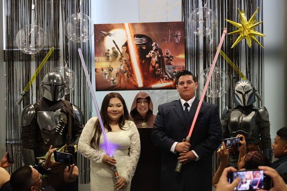 A couple in wedding attire holds lightsabers, surrounded by people in sci-fi costumes. A "Star Wars" poster and decorations in the background.